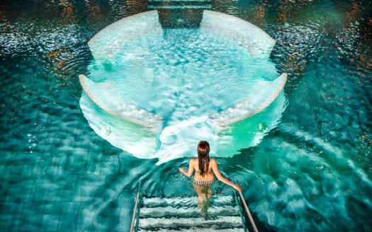 Frau steigt über eine helle Stiegen in ein Wasserbecken mit klarem türkisem Wasser. In der Mitte ist ein runder Sitzbereich. Asia Hotel & Spa