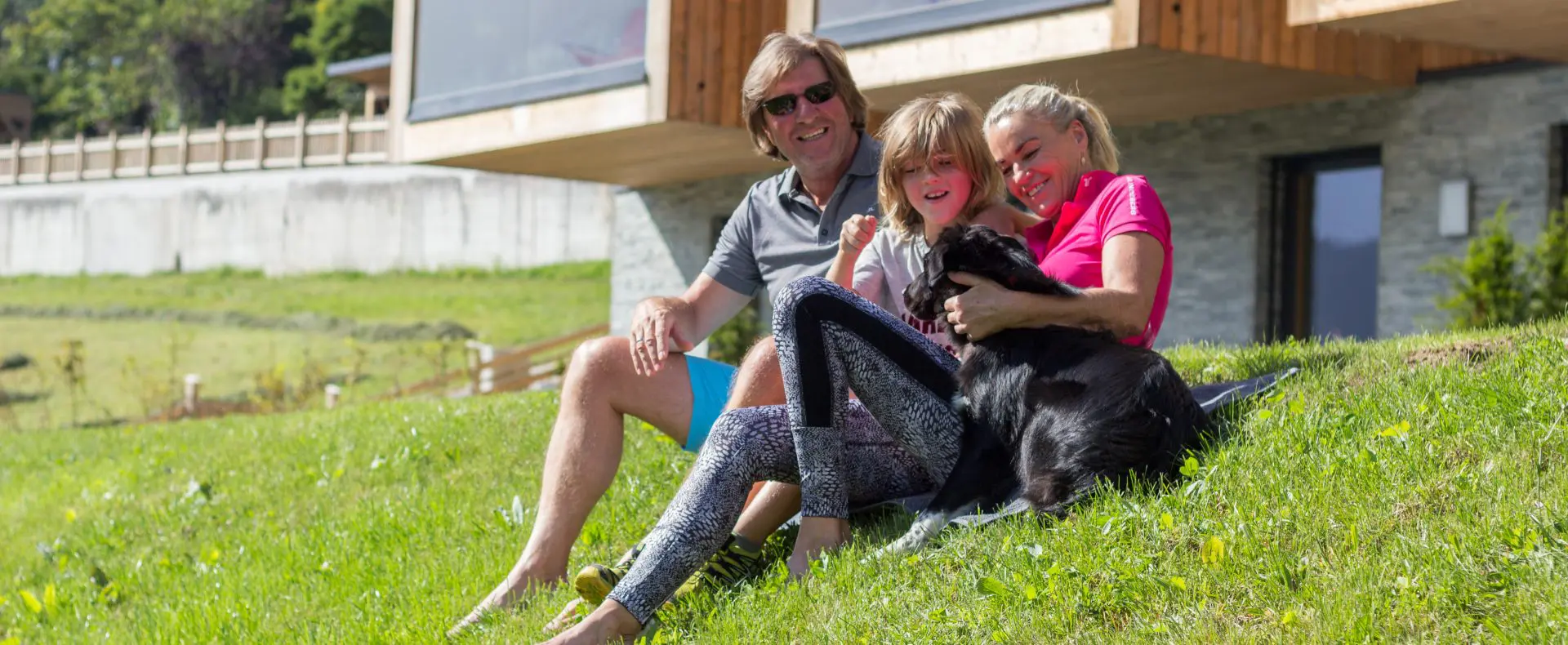 Drei Personen sitzen mit einem schwarzen Hund auf der Wiese vor der Sun Lodge.