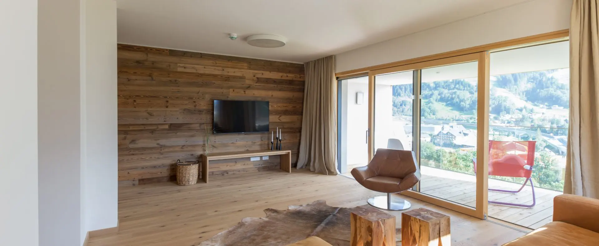 Zimmer in der Sun Lodge, eingerichtet mit einem Fernseher, einem Sessel, kleinem Tisch und Sofa. Die rechte Seite besteht aus einer Glasfront mit dahinterliegendem Balkon.