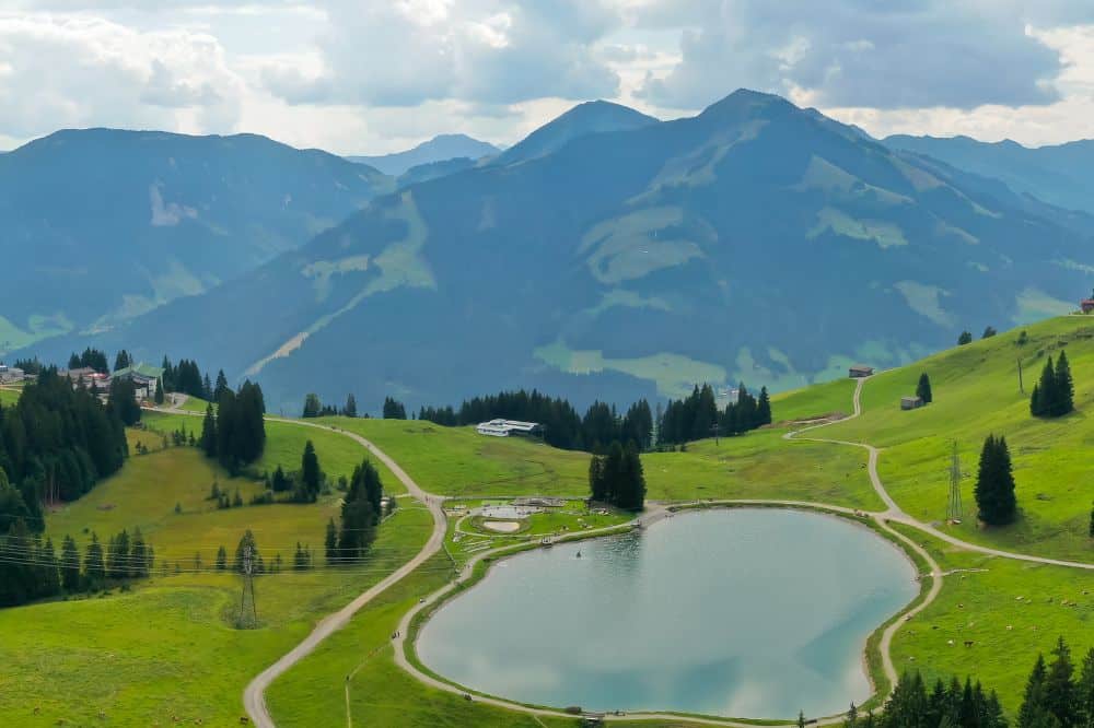 Ein ruhiger Bergsee, umgeben von grünen Wiesen und dichten Wäldern, mit hohen Bergen in der Ferne unter einem bewölkten Himmel.