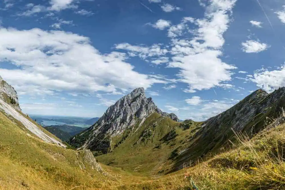Ein atemberaubender Blick auf die schroffen Gipfel der Kitzbüheler Alpen unter einem klaren, blauen Himmel. Die Wanderwege schlängeln sich durch grüne Täler, während sich in der Ferne ein See erstreckt.