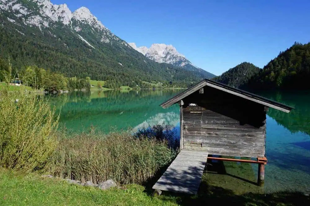 Eine kleine Holzhütte am Ufer eines kristallklaren Sees, umgeben von den majestätischen Bergen der Kitzbüheler Alpen. Das Wasser spiegelt die malerische Landschaft wider.