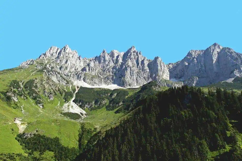 Die imposanten Gipfel des Wilden Kaisers ragen schroff und majestätisch in den strahlend blauen Himmel, während grüne Wälder die unteren Hänge bedecken.