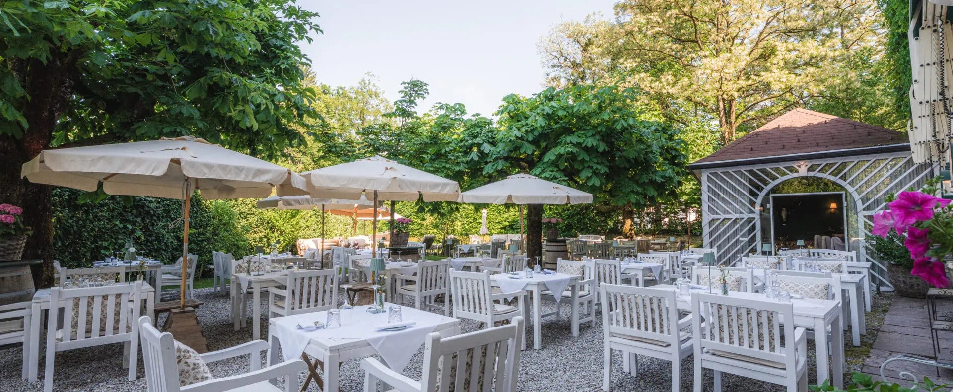 Gastgarten des Schlosswirt zu Anif mit weißen Tischen, Sonnenschirmen und viel Grün.