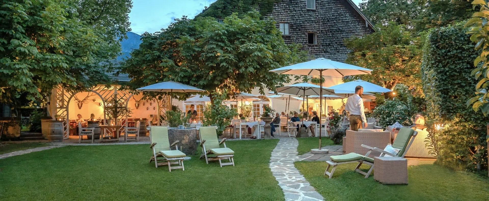 Stimmungsvoller Gastgarten des Schlosswirt zu Anif am Abend mit gedeckten Tischen, Sonnenschirmen und grünem Garten.