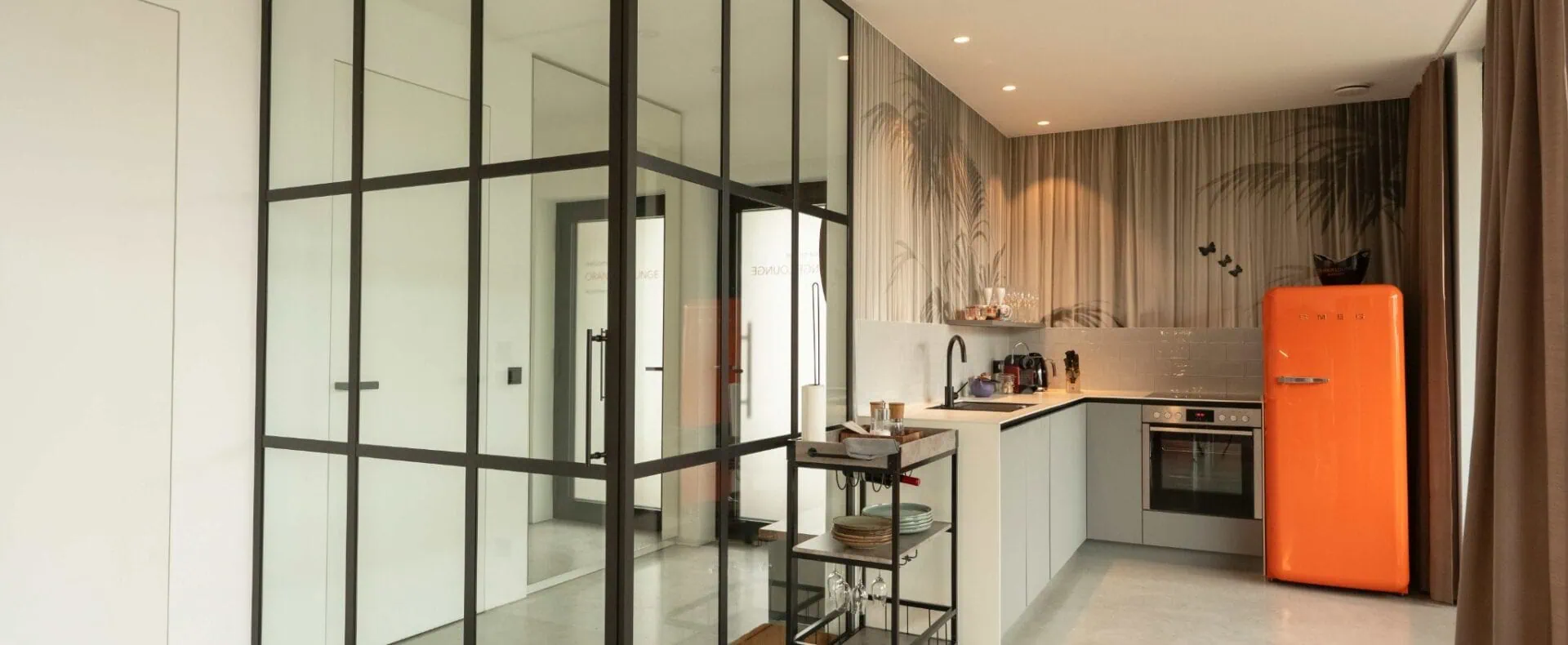 Offener Küchenbereich im Design-Apartment der ORANGE LOUNGE am Attersee mit Glastrennwand und orangefarbenem Kühlschrank.