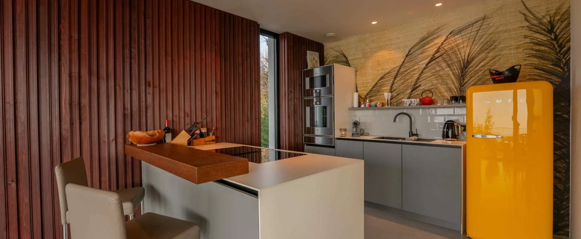 Moderne Küche der ORANGE LOUNGE am Attersee mit Kochinsel, Holzverkleidung und auffälligem gelbem Kühlschrank.